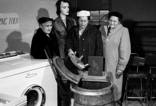 Mrs. Herman Erfurth (Verona), Mrs. Edgar Messer (Beaver Dam), and Mrs. LaVerne Johnson (Verona). There is a mannequin, (in the background) and women are viewing an exhibit of old and new laundry equipment at Farm and Home Week.