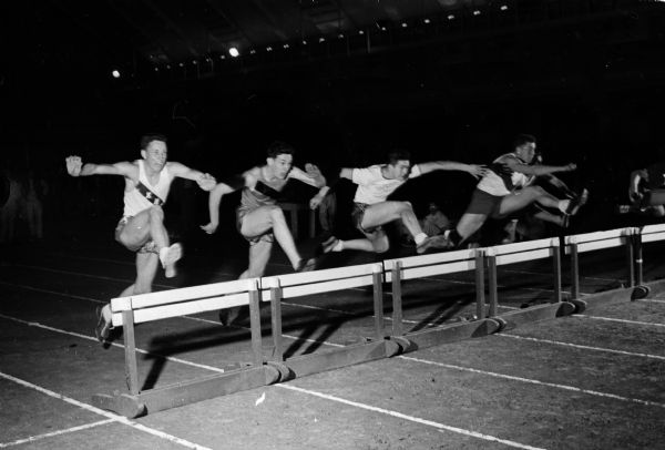 Action shot of the Madison West Relays Class B 70-yard low hurdles final race. Competitors include, from left to right: Darrel Jansen, Kimberly; Karl Hecker, Plymouth; Ron Kertacher, Port Washington, and Ted Harris, Mineral Point. Harris won the race.