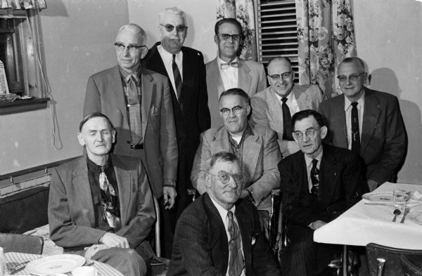 Group portrait of nine members of the Mt. Horeb Rifle Club at their banquet meeting.  In the back row, L-R, are: Elmer Culver, Clarence Lund, Bert Frederickson, the Rev. Hector Gunderson, and Red Thousand. In the middle row, seated, are: Walter Hanson, Ted Church, and Ollie Statz. Seated front and center, is: Forrest (Pops) Henderson, age 62, a long time member of the club, and a 30 year competitive marksman.