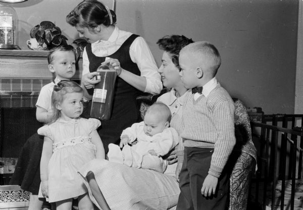 The 1958 red measles season has started in Madison. Marilyn Kelso, her son Robbie Kelso, seated on her lap, and son Tom Kelso, on the right, received gamma globulin injections to minimize the effects of the red measles. On the left are Mike Bartlett and Mary Bartlett who also received gamma globulin injections which minimized the effects of the red measles. Phyllis Bartlett is shown holding a Red Cross plasma pool from which gamma globulin can be processed.