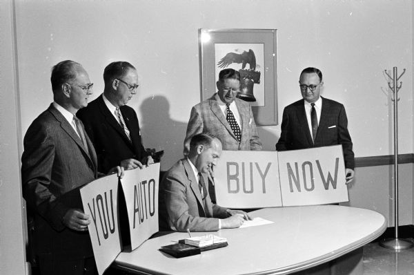 Four auto dealers are holding four signs saying "You," "Auto," "Buy," "Now." A fifth seated man, Mayor Ivan Nestingen, signs a proclamation designating, "Auto Buy Week." The men standing are: C.L. (Duke) Duquaine (Gillespie-Blumer Motors); Vern Giesler (Schappe Motors Inc.); Warner Young (Young's Studebaker Sales); and Clarence Thorstad (Hult's Capital Garage).