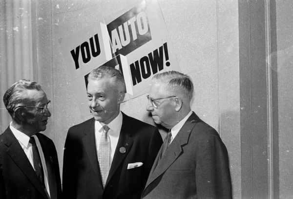 Two Madison automobile dealers confer with B.R. Durkee (center), director of advertising and sales promotion, Chrysler Corp. division. At left is J.P. Gillespie (Gillespie-Blumer Motors, Inc.). At right is Ralph Hult (Hult's Capital Garage). A sign on the wall proclaims: "You Auto Buy Now."