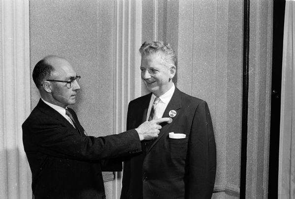 One man is pointing his index finger under a pin on the lapel of another man. The pin says: "Business Is Good." The pin is worn by Melvin Larson, State Motor Vehicle Department commissioner. The man pointing is Dean Waters, left, of Waters Motor Co.