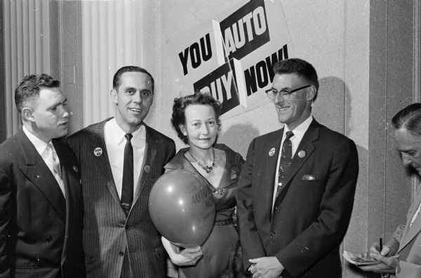 Four people posing in front of a "You Auto Buy Now" sign. Left to right, they are, Richard Young of Warner Young Co.; Kenneth Kimport, Kayser Motors; Julia Kimport, who is holding an inflated balloon; and Ray Olson of the Rockford district, Ford Motor Co.