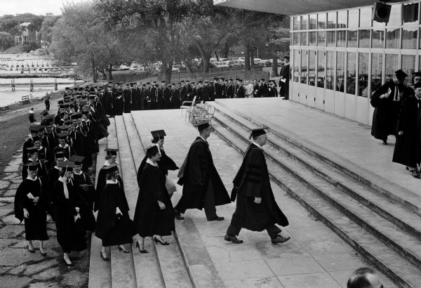 Elevated view of a long line of University of Wisconsin honor students in caps and gowns parading across Union Terrace and up the steps into the Union Theater to attend the University Honors Convocation.