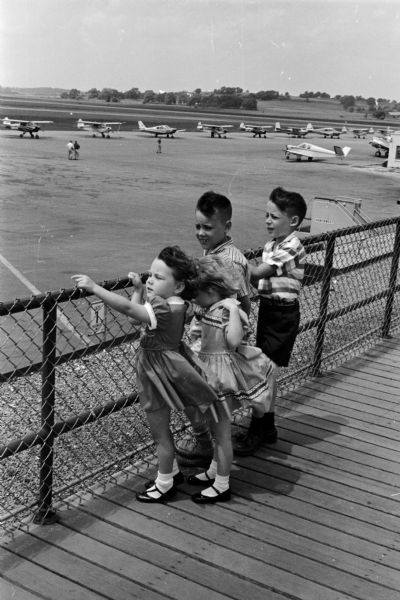 Four children stand on the observation deck watching planes at the Madison Municipal airport. The children include, from left to right: Carol, 3, Christine, 4, Michael, 8, and Stephen, 9. Their parents are Mr. and Mrs. Richard Usher of Montgomery, Alabama. The photograph was taken as part of an article suggesting Madison places and activities for summertime visiting.