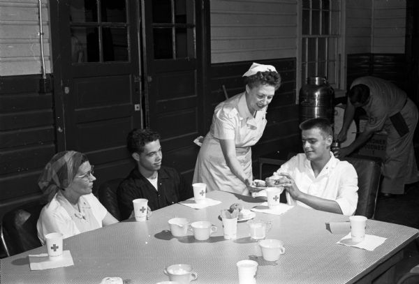 Helen Thomson, wife of the governor, is among the Red Cross canteen volunteer hostesses who serve coffee and doughnuts to Truax airmen giving blood at the Service Club. Being served are Mrs. Donald Stilwell, substituting for her sergeant husband who was ineligible, Azores-born A 2/C Everisto DaSilva, now of New York City, and A 2/C Frank Kahre, from Vincennes, Indiana.