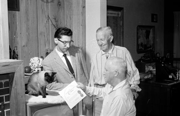 Charles B. Seals, and his father Bellamy, have received a citation from the Puss'n Boots bronze award committee. The citation resulted from pictures of their Siamese cat, Thai, ringing a doorbell at their home at 1213 Wingra Drive. The pictures, taken by Edwin Stein and printed in an April 27, 1958 "We Saw You" feature in the Wisconsin State Journal, show how Thai can summon one of her owners to the door by pressing a low-level bell made by Charles. The owners' award was presented on behalf of Thai, who was assisted in the ceremony by Edwin Stein, State Journal photographer, who took the pictures for the feature. Shown admiring the citation, are, left to right, Thai, Ed Stein, Bellamy Seals, and seated, Charles Seals.