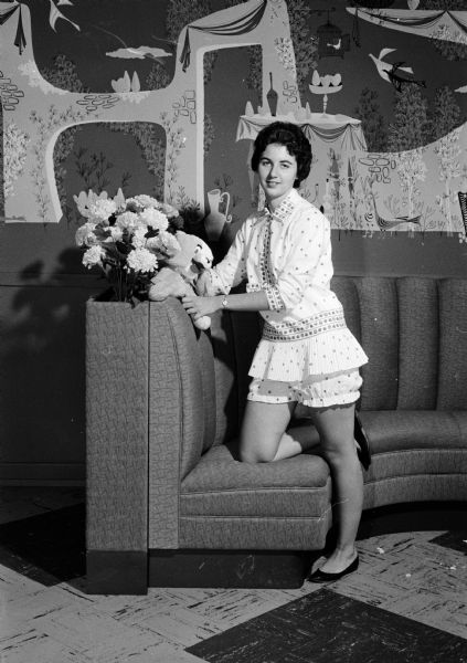 Jane Hansen leans against a sofa while modeling a two-piece baby doll pajama set in a chemise style.