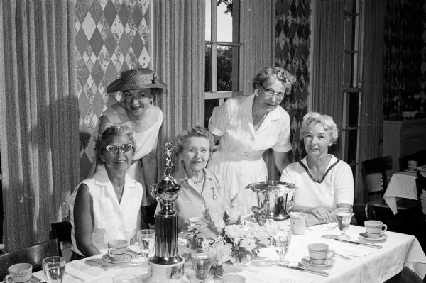 Group portrait of those gathered at the head table during a luncheon that accompanied the Madison Women's Interclub golf play. Attendees include, from left to right: Dorothy Steiro, Blackhawk Interclub chairman; Selma Schubring, Maple Bluff; and Mable Day, Nakoma Interclub chairman. Standing are Marion Pecher, and Eleanor Persons, Maple Bluff chairman. Miss Pecher, a Blackhawk member presented the Beecroft trophy, for handicap play, for her uncle Dr. Beecroft. The Blackhawk women will retain the Beecroft trophy permanently, having won it for the third consecutive year. The Schubring trophy, for scratch play, is also on the table.