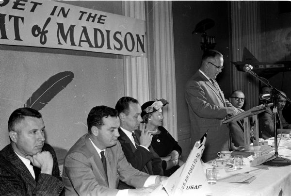 Ted Meloy (second from right), drive chairman, stands at a podium while informing the group that they have obtained over half their goal. Listening, left to right, are: John Brickhouse, area business chairman; Nicholas Lesselyoung, government chairman; and Constance Holmquist, women's division chairman. At far right is R.A. (Hans) Eissfeldt, president of United Givers. On their table is a large scale-model U.S. air force jet.
