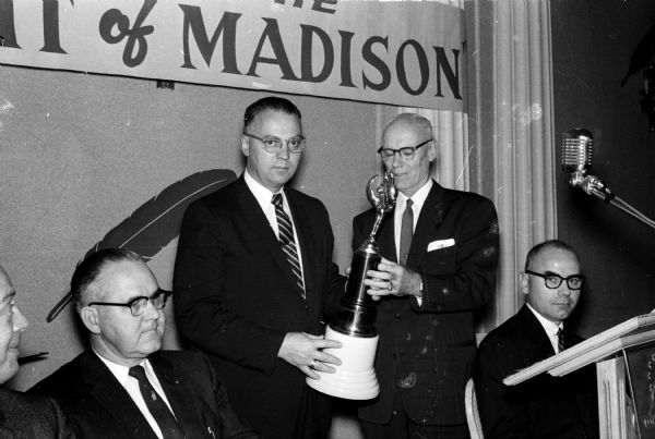 The Madison Chamber of Commerce trophy for the division of the United Givers' Fund achieving the highest percentage of its goal is awarded to the business and industry division at the fund victory dinner. Shown from left to right are: Theodore M. Meloy, general chairman of the United Givers' Fund campaign; Wilburt Wittwer, division chairman; and Marshall Browne, Chamber of Commerce president. Man seated on the right is unidentified.