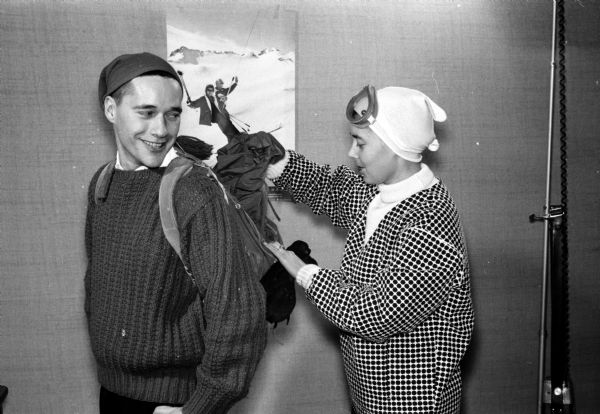 Anne Collins packs a parka into the knapsack worn by her husband Edward Collins at a fashion show for the Madison YWCA Ski Club. Both are modeling ski apparel. Mr. Collins is vice president of the YMCA Ski Club, and is also its head instructor.