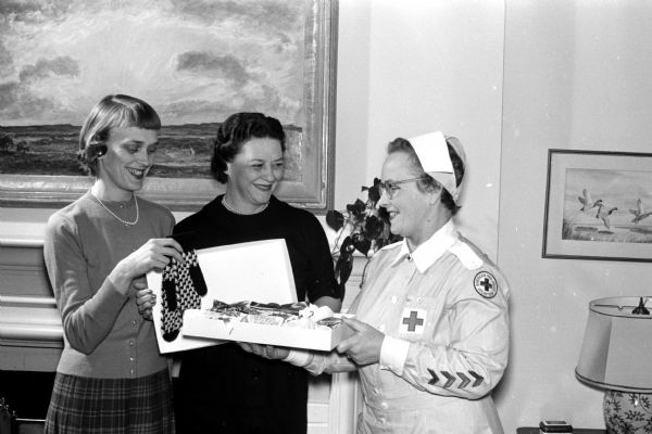 Three women pack Christmas gifts as part of a project in which members of 30 Dane County organizations "adopt" a patient at Mendota State Hospital. Participants include, from left to right: Caroline Worcester, president of Alpha Omicron Pi alumnae;  Theadora Diener, alumnae vice-president; and Mrs. A.M. Anderson, chairman of the Red Cross Gray Lady organization. They are preparing a package of warm mittens for 25 adopted women patients.