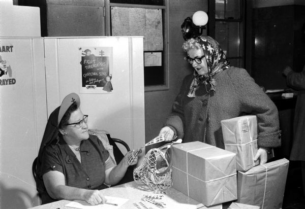 Esther Haack (seated left) sells a page of Christmas seals to Leone Wagner at the post office. The Madison Tuberculosis Association sells about $150 worth of seals at the post office each year.