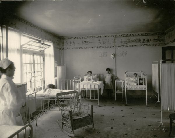 Light from a large window brightens the children's ward at Madison General Hospital.  A nurse in cap and uniform stands in the left foreground.  A young girl stands against the far wall between two iron patient beds; a child sits up in each of the two beds. Other furnishings include a wicker rocker, privacy screens, and bedside tables. The floor is tiled. There is a painted or stenciled frieze around the high-ceilinged room depicting windmills, sailboats and Dutch children at play.