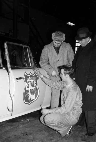 Mike Gilomen, an employee of Capital Auto Body, applies a state patrol decal to the side of the first American Motors Ambassador to be added to the patrol's fleet. All of the patrol's 1956 models and some of the 1957 models are being replaced with 53 Ambassadors and 46 Fords. Looking on are Norbert Anderson, left, of the State Motor Vehicle Department, and Ed Moore, Kenosha, of American Motors Corporation.