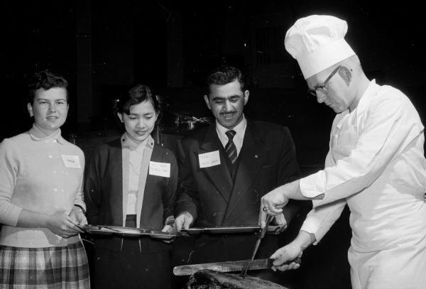 The Madison Rotary Club members honor 150 foreign students attending the University of Wisconsin at a fiesta at the University of Wisconsin Stock Pavilion. Tom Weth carves the roast for three of the guests. They are, left to right: Rose Marie Herschmann, Chili; Thi Hein Ngo, Vietnam; and Seleh Sadig, Iraq.