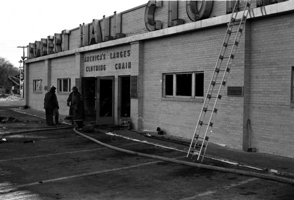 View of the front of a fire-damaged commercial building at 3205 East Washington Avenue with two fire fighters, two hoses, and a ladder nearby. The signage on top of the building reads, "Robert Hall Clothing." The signage above the doorway says, "America's Largest Clothing Chain."