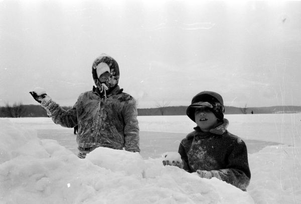 Winter scene with brothers Greg and Mark Hennen of 5709 Midmoor Road standing in their snow fort and holding snowballs at the ready the day following a five-inch snowfall. Behind them is a large open field in front of a forested horizon with a tall hill behind.