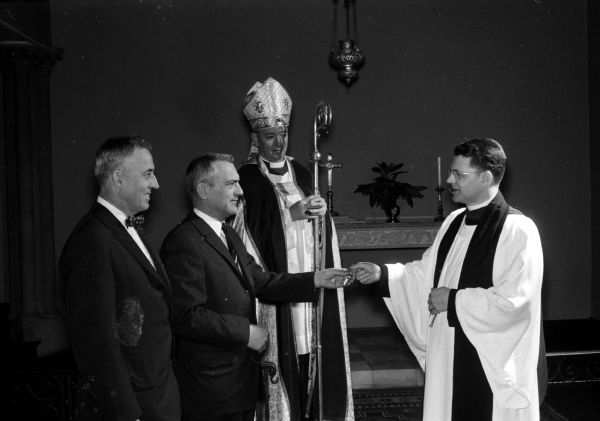 The Rev. Fr. Paul Z. Hoornstra (right) receives the keys to the church from Senior Warden Charles Burdick (second from left). At left is Junior Warden J. Nash Williams. At the rear, with miter, vestments and staff (crosier), is the Rt. Rev. Donald Hallock, Episcopal bishop of the Milwaukee diocese, who conducted the installation. Grace Episcopal Church is on the Capitol Square at 6 North Carroll Street.