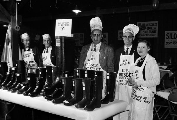 Four men and one woman stand behind a table displaying rubber boots.  They are each wearing a tall white hat with the words: "I'm A Butter Cook" and an apron with the person's name, the company they represent, and the words "You have served us, let us serve you."  
Persons and the company names are, left to right: Clarence Fenske, Madison Dairy Supply; Verne Miller, Madison Dairy Supply; Al Negus, president of the  Madison Dairy Supply; Len Mueller, U.S. Rubber Co.; and Marcella Negus, secretary-treasurer of Madison Dairy Supply Co.