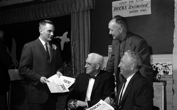 420 men turned out for the annual Ducks Unlimited dinner at Club Chanticleer.  Ducks Unlimited was founded in 1938 by persons interested in wildlife conservation. Thomas Towell, left, hands out copies of the winter issue of the quarterly magazine of the organization. Seated, left to right, are: Dr. E.F. Johnson and E.H. Fisher. Standing at right is B.L. Mackin, Poynette.