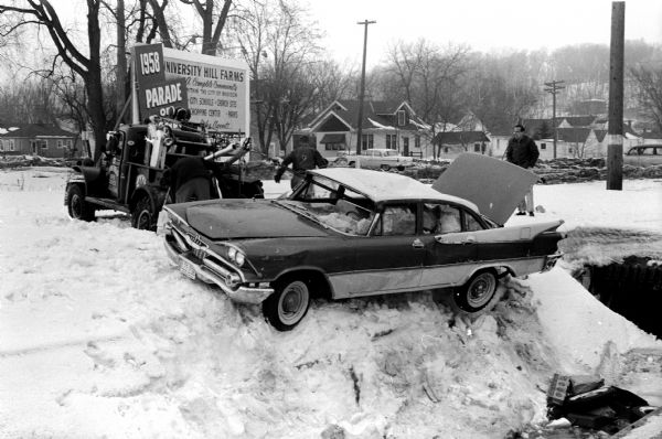 Winter scene with garagemen retrieving another car from the open drainage ditch on Midvale Boulevard. The 1959 Dodge skidded into a snowbank and catapulted 12 feet in the air, landing upside down in the ditch. Occupants were treated at a local hospital. Sign in background announces 1958 Parade of Homes in the University Hill Farms neighborhood.