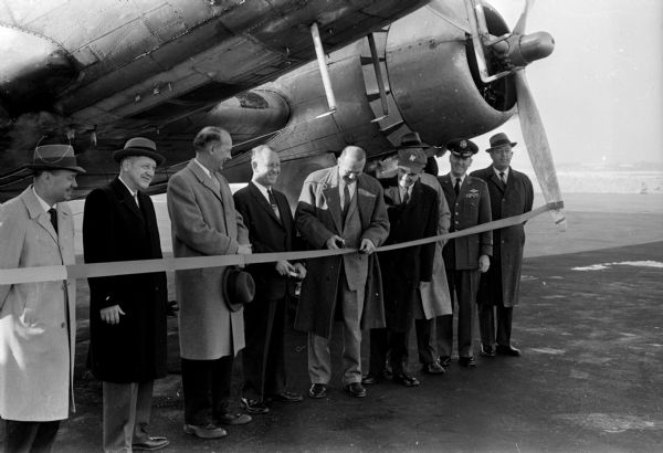 A group of men posing in front of an engine and under the fuselage of a DC-3 air liner. The original caption states: "Cutting the tape for the inaugural flight of Ozark Air Lines in Madison is Secretary of State Robert Zimmerman. On hand to greet the DC-3 at Municipal Airport were, left to right: Robert Skuldt, manager of the airport; Stan W. Orr, president of the Madison Chamber of Commerce; Mayor Ivan Nestingen; Laddie Hamilton, president of Ozark; Zimmerman; Ernest Harr, chairman of the chamber's aviation committee; Col. John Pease, commander at Truax Field; and I. W. Lackore, executive director of the Chamber of Commerce."