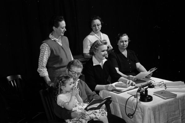 Six women, three with scripts, rehearse at a table set with a prop telephone. The original caption states: "Members of St. James Players will present a playlet, Mrs. Zany's Problems," at the spring meeting of the Madison Deanery of the Diocesan Council of Catholic Women at St. Raphael Cathedral." The players shown include Joan McWilliams (a little girl), Agnes Brinkman, Roselyn McWilliams, and Mary Haas. Seated are Lydia Coyne and Agnes Rapp.