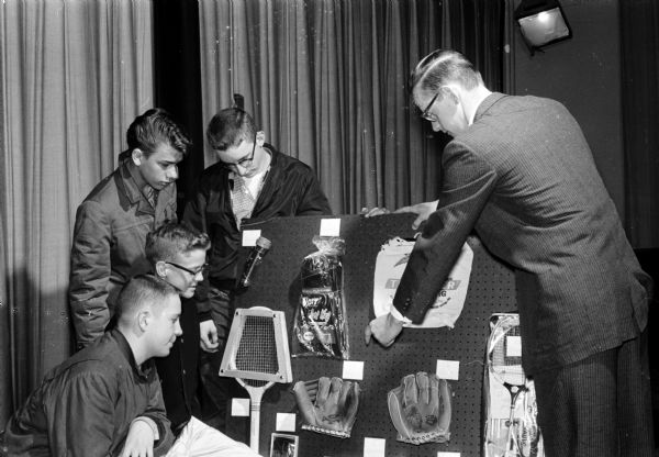 Four Madison Newspapers, Inc., carrier salesmen (or newspaper home delivery boys) looking over possible awards for new subscriptions, such as prizes, cash, or trips to Chicago. District Manager Louis Dahl (right) is pointing at some prizes. Ken Cole and Bill Rogers are in the foreground, while Don Dotzel and Dave McCaffrey are standing in back.