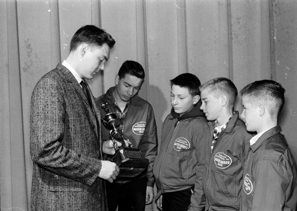 Four Madison Newspapers, Inc., carrier salesmen (or newspaper home delivery boys) looking over possible awards for new subscriptions, such as prizes, cash, or trips to Chicago. District Manager Robert Snelling, (right) is showing them a trophy. The four boys are wearing identical jackets with patches indicating they are outstanding carriers. The boys names are Craig Rennebohm, George Scott, Jerry Meisteer, and Bill Kuehn.