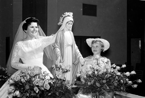 A crown of roses and lilies-of-the-valley are placed on a statue of the Virgin Mary during Edgewood College's May Crowning ceremonies. Mary Jane Roach, on the left, is wearing a white bridal dress in her role as crowner. At right is Claire Hunold who carried the crown during the ceremony in St. Joseph's Chapel.