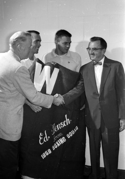 Members of the 1959 University of Wisconsin boxing team present Eddie Gensch with a W blanket in recognition of his contributions to the boxing team. Gensch has been an unofficial manager and cheerleader of Wisconsin boxing teams for several years. Shown presenting the blanket (left to right) are Coach Vern Woodward; Bill Sensiha, co-captain elect of the 1960 boxing team; Bob Christopherson, co-captain of the 1959 boxing team; and Gensch.