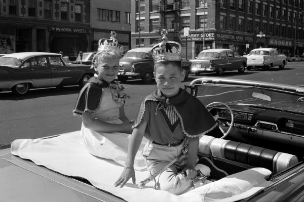A girl and a boy, both wearing crowns, are posing on the open back of a convertible top automobile. They are the queen and king of the Bicycle Safety Week parade, Jean Dauck and Jon Miyagawa.