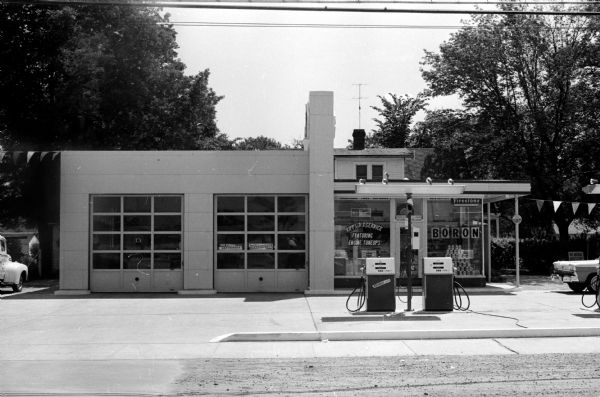 View of a Mid Century-style service station with two service bays and a sales room.  Three gas pumps and the trunk of a car are visible and trees stand in the background.