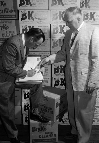Two men pose by a wall of highly stacked boxes, each marked with a large "BK" logo. One of the men holds a pen and is bent over a notebook.