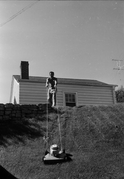 Richard Brachman, an engineer, demonstrates his solution for the problem of mowing the grass on a steep slope near his home. He removed the handles of his power mower, attached a pair of ropes, and now steers the mower as he pulls it up the grade and lets it coast back down.