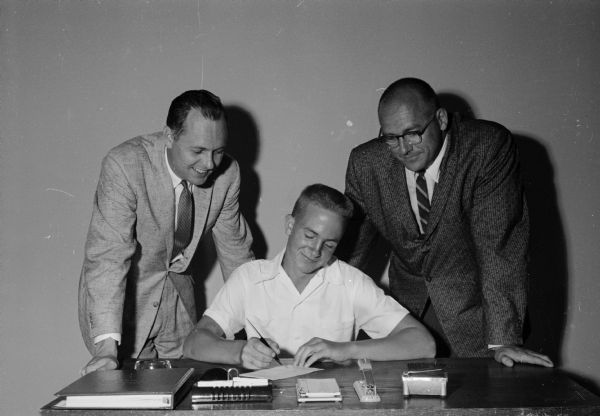 Loren Thompson, seated, becomes the first teenager to become a member of the Madison Junior Achievement Corporation. Looking on are Don Alexander, secretary and public relations director of Red Dot Foods, Inc.; and president John T. Lunenschloss, owner-operator of Lunenschloss Sporting Goods store and associated with Madison-Kipp Corporation.
