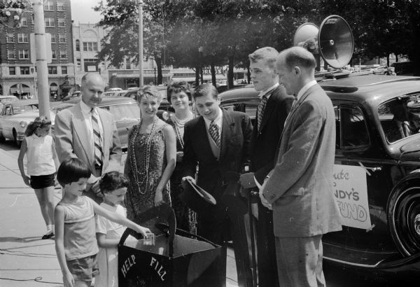 Martha and Betsy Dapin, left, making the first contribution on Capitol Square for the Roundy's Fun Fund. Standing nearby is Atty. James Geisler, chair of the Fun Fund committee; Claudia Traisman, Mary Coogan, Jeffrey Kravat, Jeffrey Culbertson and Mayor Ivan A. Nestingen. In the background is an early 1930's model Packard car and two of the youth are wearing 1920 fashions. The fund is used to aid handicapped children.