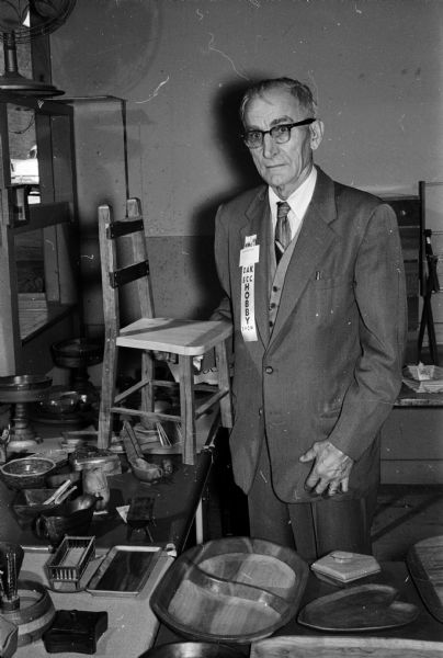John Uselman standing with a chair he made out of scraps of wood at the hobby and talent show at the Madison Community Center. The one-day show was sponsored by the Older Adult Klub (OAK) and senior citizen clubs. Unpublished photographs of a man holding a weaving and of W.F. Schroeder who records weather information with his instruments are in the series.