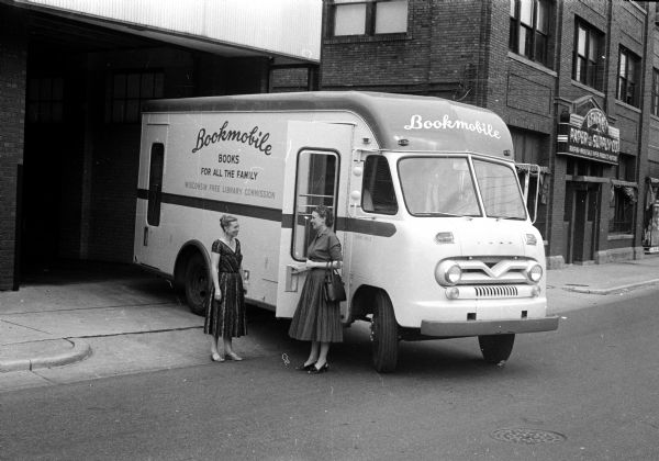 Ione Nelson, left and S. Janice Kee are standing next to the Wisconsin Free Library commission's new bookmobile. Ione Nelson is the public library consultant in charge of the project, and Janice Kee is secretary of the commission. The building in the background is the General Paper and Supply Company at 714 Williamson Street.