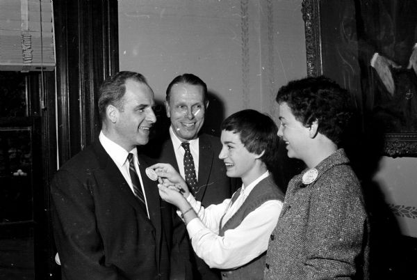 U.W. student Betty Miller pins a homecoming button on Governor Gaylord Nelson's lapel with U.W. President Conrad Elvejem looking on. At right is U.W. student Kitty McGinnis.