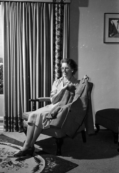 Kate Schultz is shown with a sport coat she made for her husband from fabric that she wove. She also hooked the rug at her feet, upholstered the chair she is sitting in from fabrics she wove, and block printed the draperies behind her.