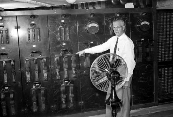 Frank E. Nordeen, a state engineering employee, pointing to the main electric switchboard in the State Capitol building basement, while standing next to an electric fan used to cool down the board.