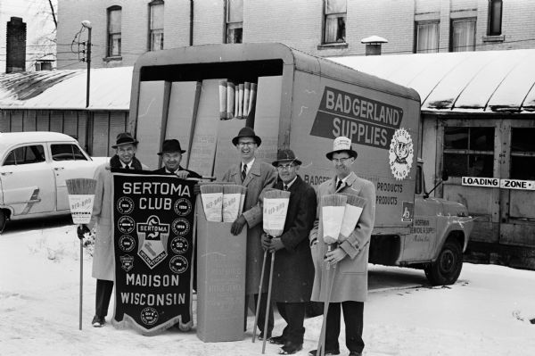 Five members of the Madison Seratoma Club, including, left to right, William Rowland, William Murphy, Harold Hueschen, Ray Sweeney, and Wendall Ward, holding brooms to advertise their upcoming project in which they hope to sell 1,000 brooms to raise funds for charitable causes. Most of the funds will be used to buy toys and other recreational equipment for children who are residents at the new Central Colony. A truck loaded with brooms is in the background.