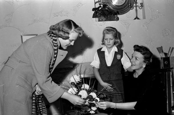 Lisa Rutherford (left), chairman of the West High Y-Teen Club and chairman of the ticket sellers for the flower show presented by the Wisconsin-Upper Michigan Florists Association, selling a floral arrangement to Elsa Johnson (right) as her young daughter, Martha Johnson (center) is looking on.