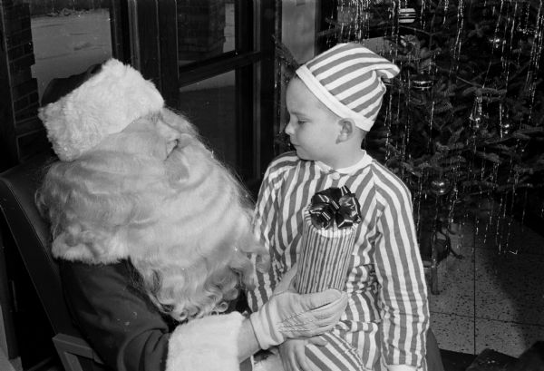 Santa presents a wrapped present in striped paper to Ricky Madsen, who is wearing striped pajamas and a striped night-cap. There is a Christmas tree in the background on the right.