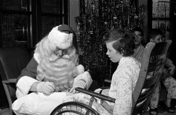 Santa autographs an above the knee full leg cast worn by Deborah Sosinsky as she looks on from her wheelchair.
