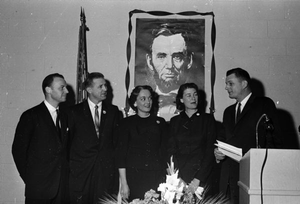 Two Republican candidates for governor and their wives chat with Roger Radue, dinner chairman, before the annual Dane County GOP Lincoln Day fund-raising dinner. A large poster of Abraham Lincoln is displayed on the wall behind them. The candidates are Phillip Kuehn, Milwaukee, and Jack Olson, Wisconsin Dells. Also shown are Mrs. Olson, Mrs. Kuehn, and Roger Radue.